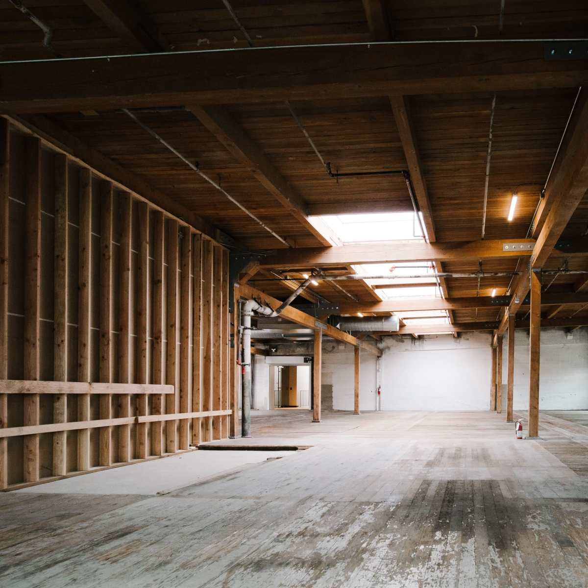 An empty industrial interior with wooden beams, large windows, and unfinished wooden flooring.