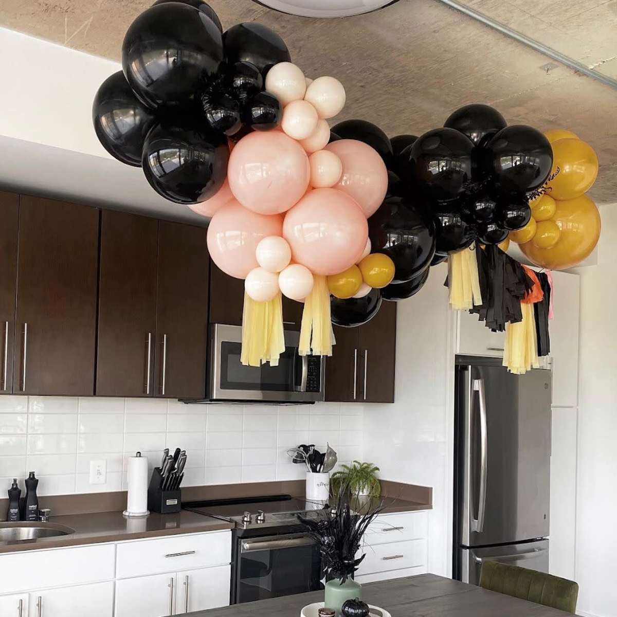 A cluster of balloons in black, pink, white, and gold hangs above a kitchen table with green chairs in a modern kitchen.