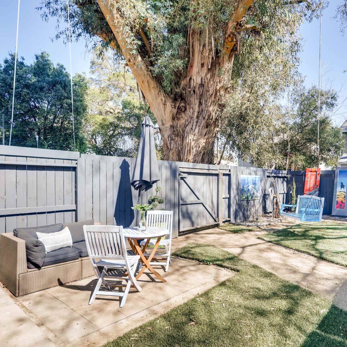 A patio area features a seating arrangement with a table and chairs, surrounded by greenery, under a large tree.