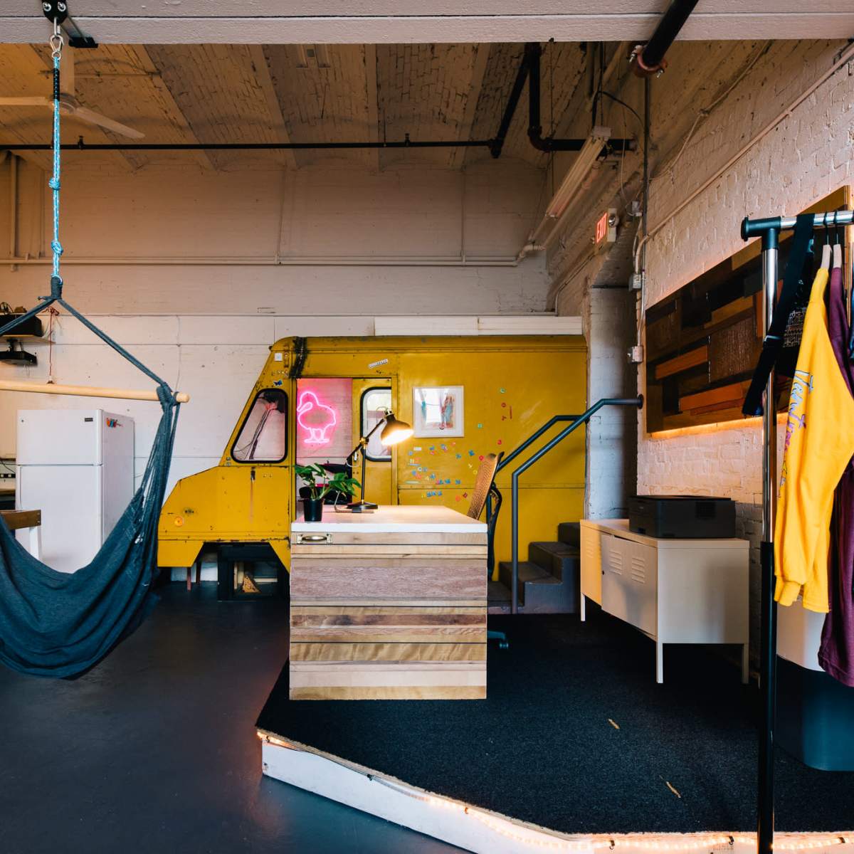 A modern interior space featuring a hammock suspended from the ceiling, a small kitchen area, and brightly colored decor including a yellow wall and neon lighting.
