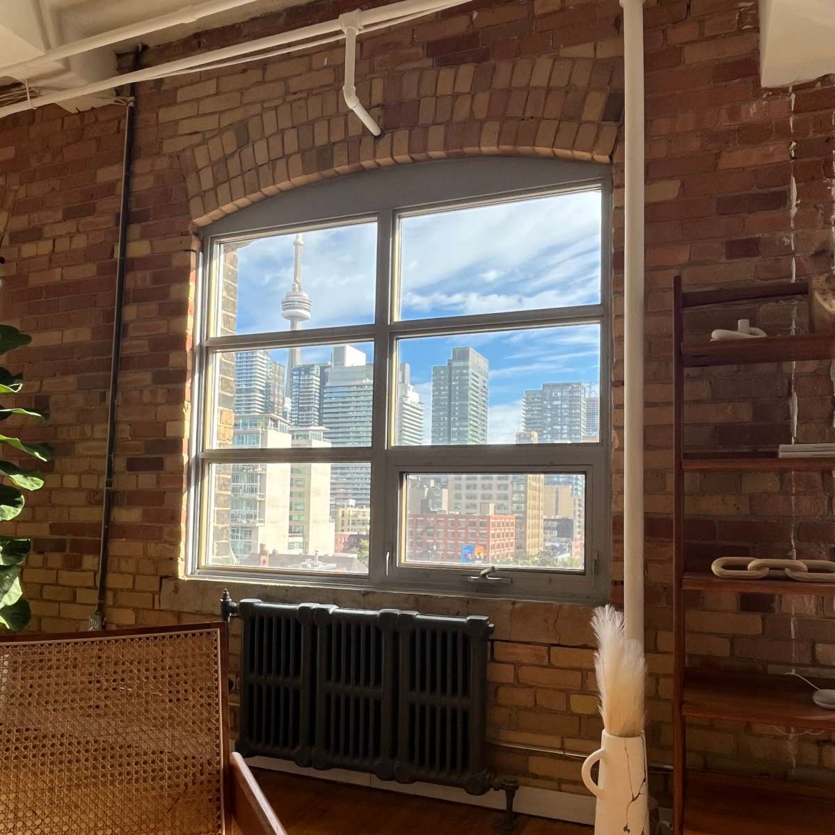 A view from a room featuring a large window that overlooks a city skyline with tall buildings, including a prominent tower, framed by a rustic brick wall.