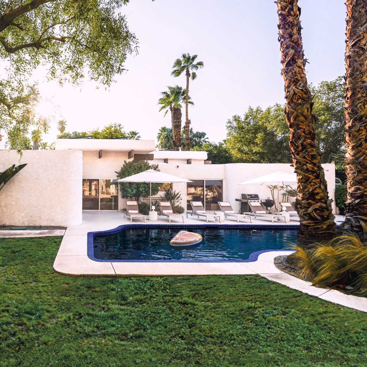 A modern home with a swimming pool and lounge chairs is situated in a landscaped yard with palm trees and tropical plants.