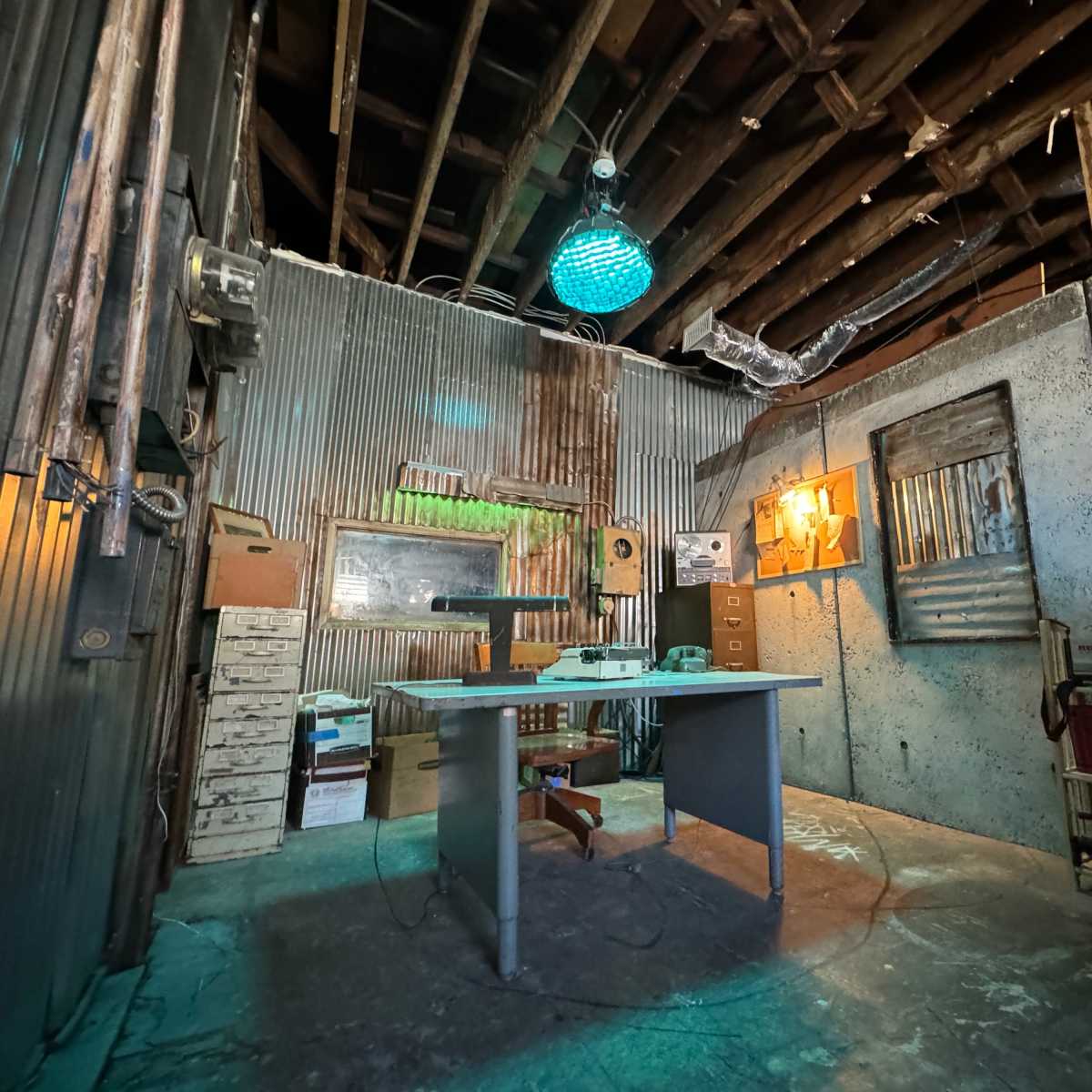 A sparsely furnished room with exposed beams, a desk at the center, and industrial-style walls featuring a green light fixture.