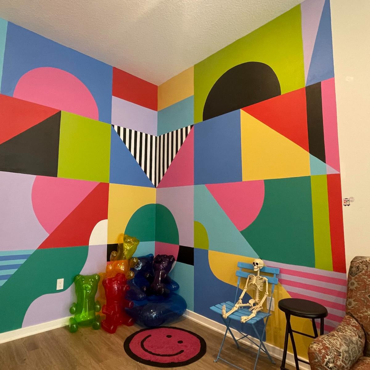 A brightly colored geometric mural on a corner wall with a skeleton, colorful plastic bears, and a smiley face rug on the floor.