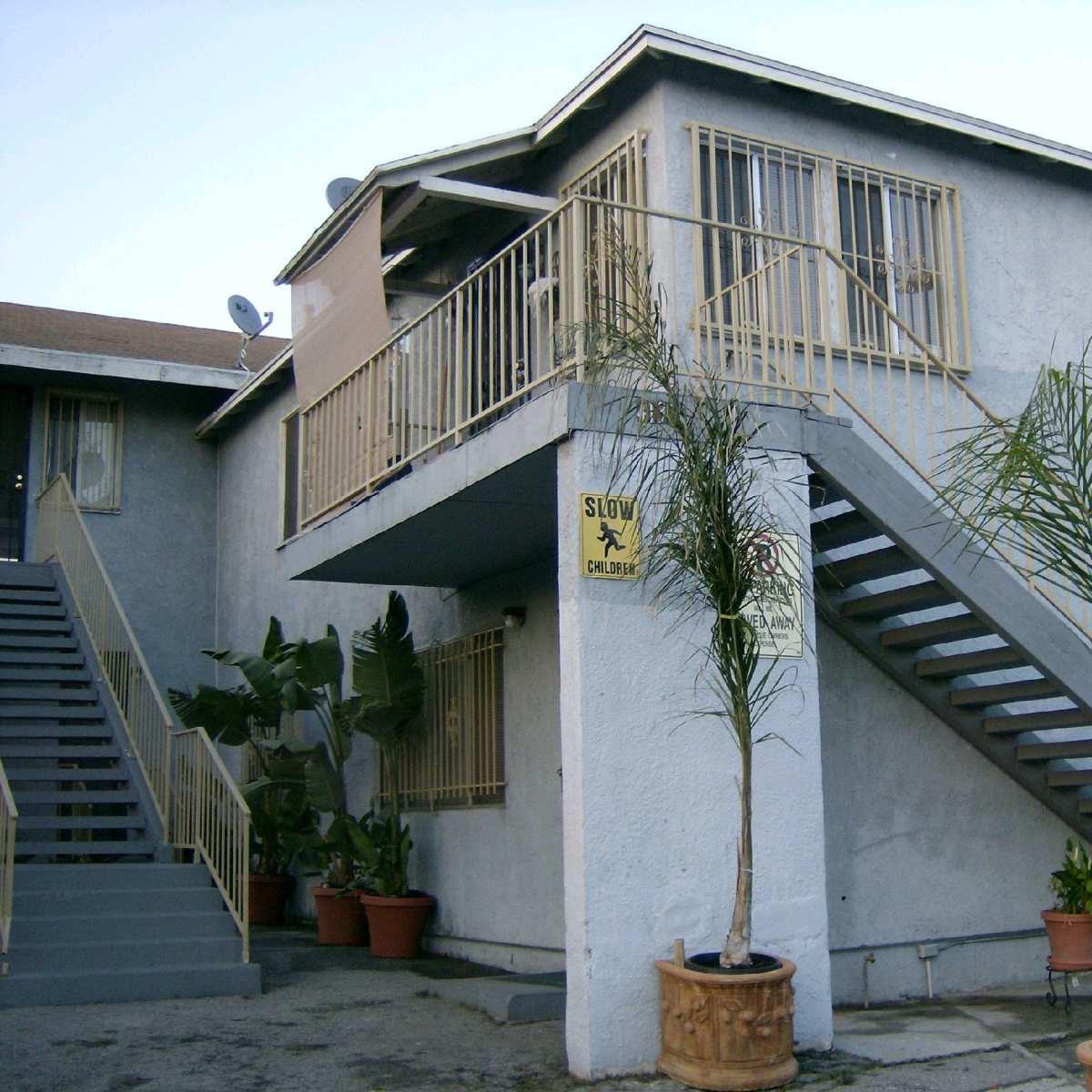 A two-story apartment building with a gray exterior, featuring a staircase on the left and potted palm plants at the entrance.