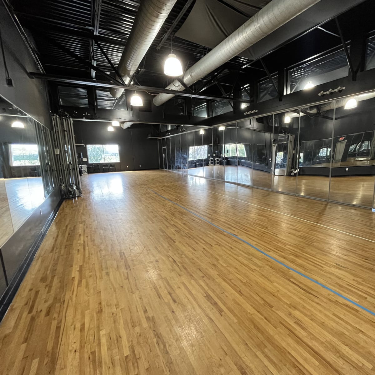 A spacious dance studio with wooden flooring, large mirrors on one wall, and bright overhead lights.