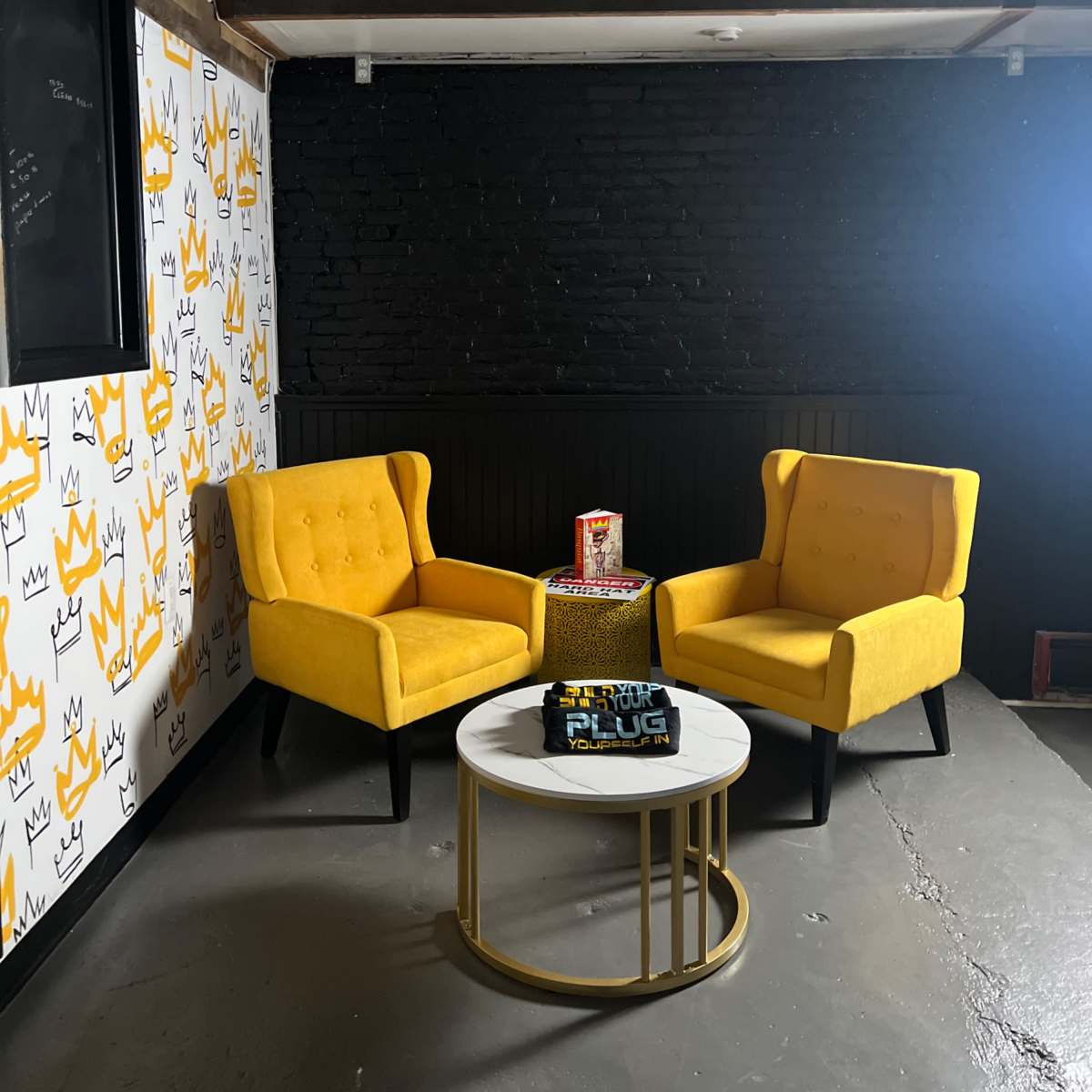 Two yellow armchairs facing a small coffee table in a room with patterned wallpaper and a dark brick wall.