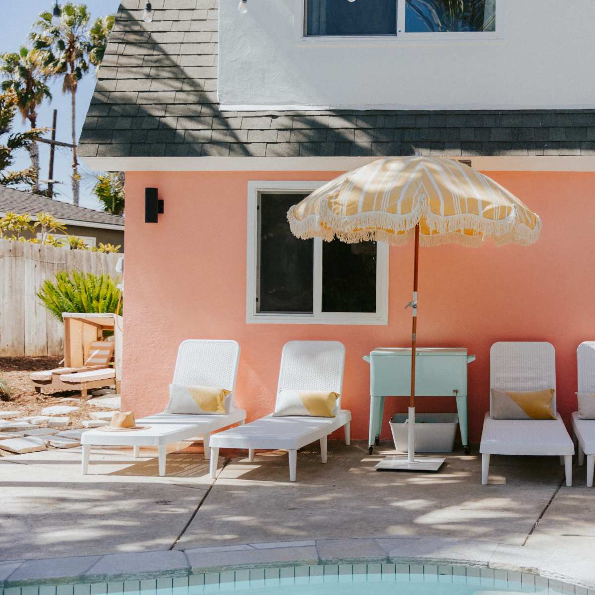 A row of white lounge chairs with yellow accent pillows sits beside a pink house and a pool, shaded by a striped umbrella.