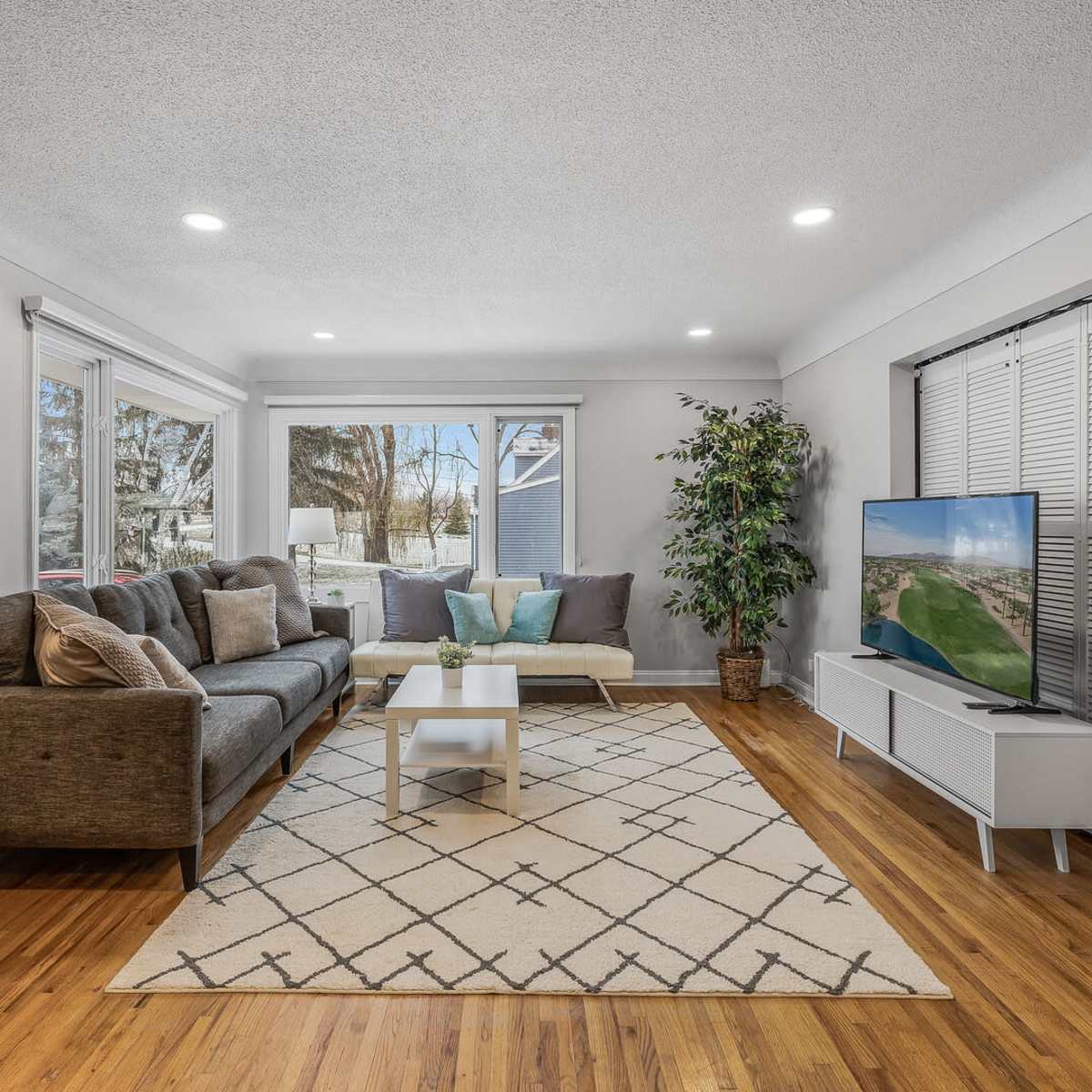 A modern living room with a brown sectional sofa, a coffee table on a geometric patterned rug, and a large television set against the wall, with windows allowing natural light to enter.