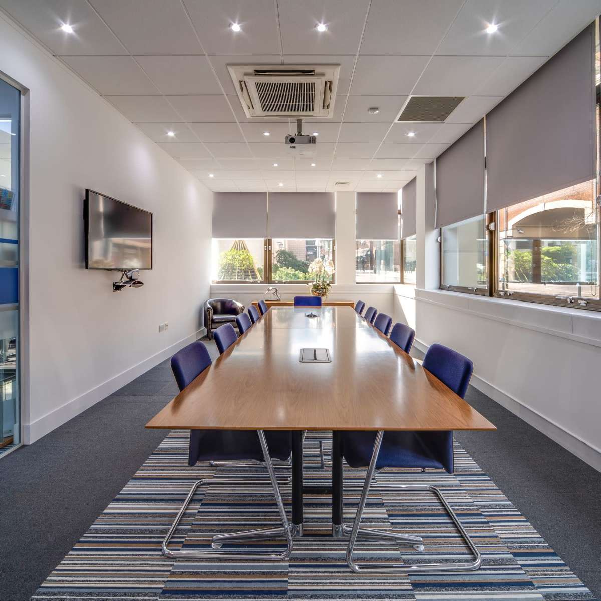 A modern conference room features a long wooden table surrounded by blue chairs, with large windows allowing natural light and views of greenery outside.