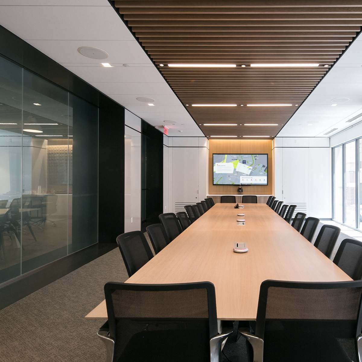 A modern conference room features a long wooden table surrounded by black mesh chairs, with large windows and a screen displaying a presentation on one wall.