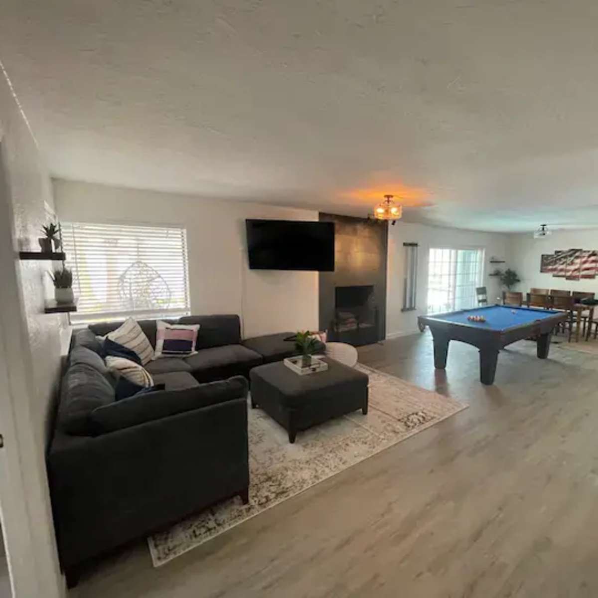 A spacious living room with a grey sectional sofa, a pool table, a dining area, and a large window providing natural light.
