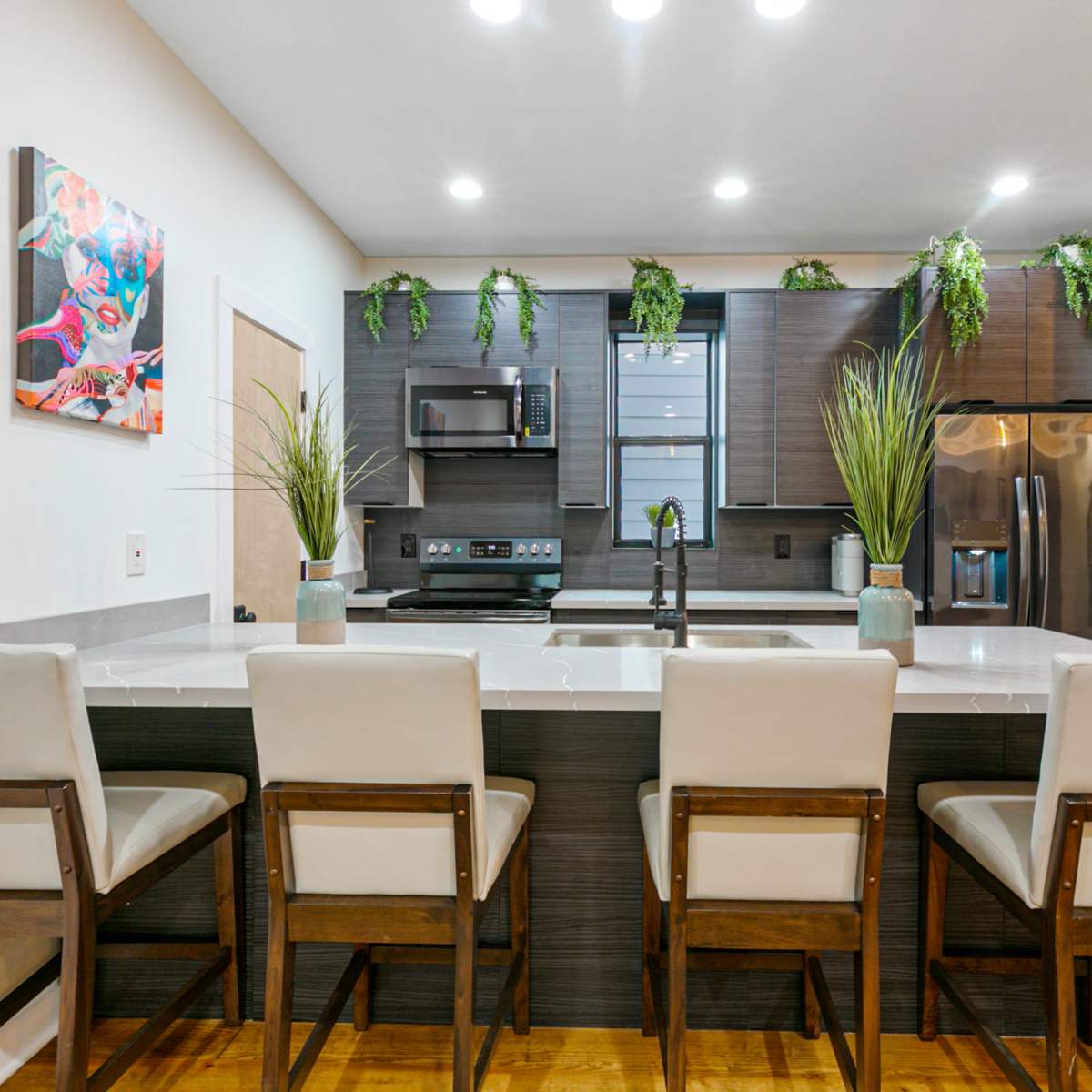 A modern kitchen features a counter with four upholstered stools, stainless steel appliances, and decorative greenery.