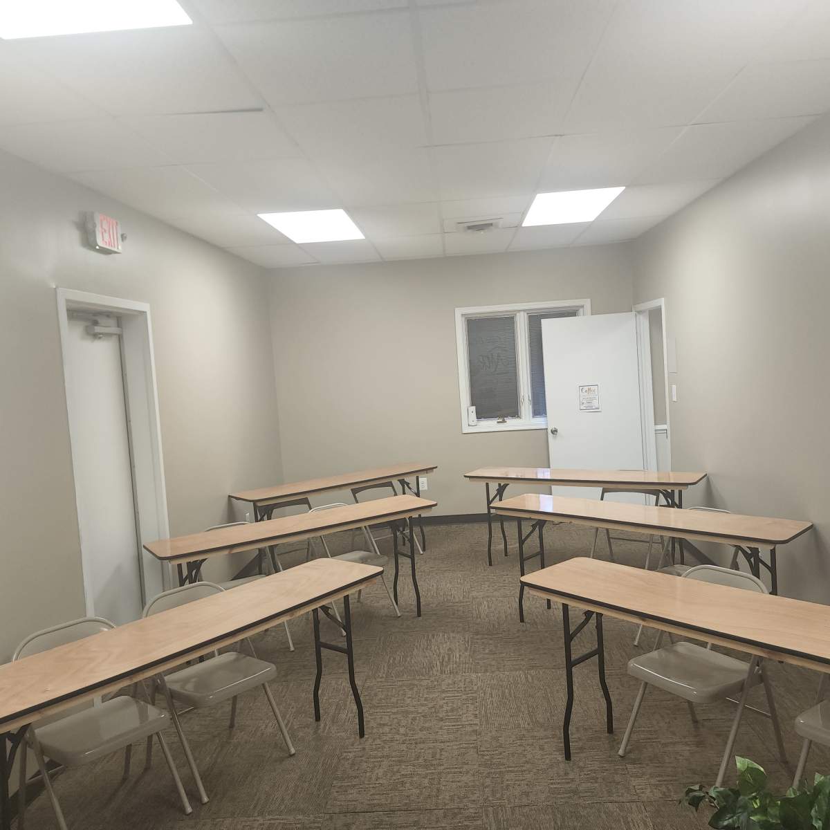 A small classroom setup with several wooden tables and folding chairs arranged in two rows in a plain, softly lit room.