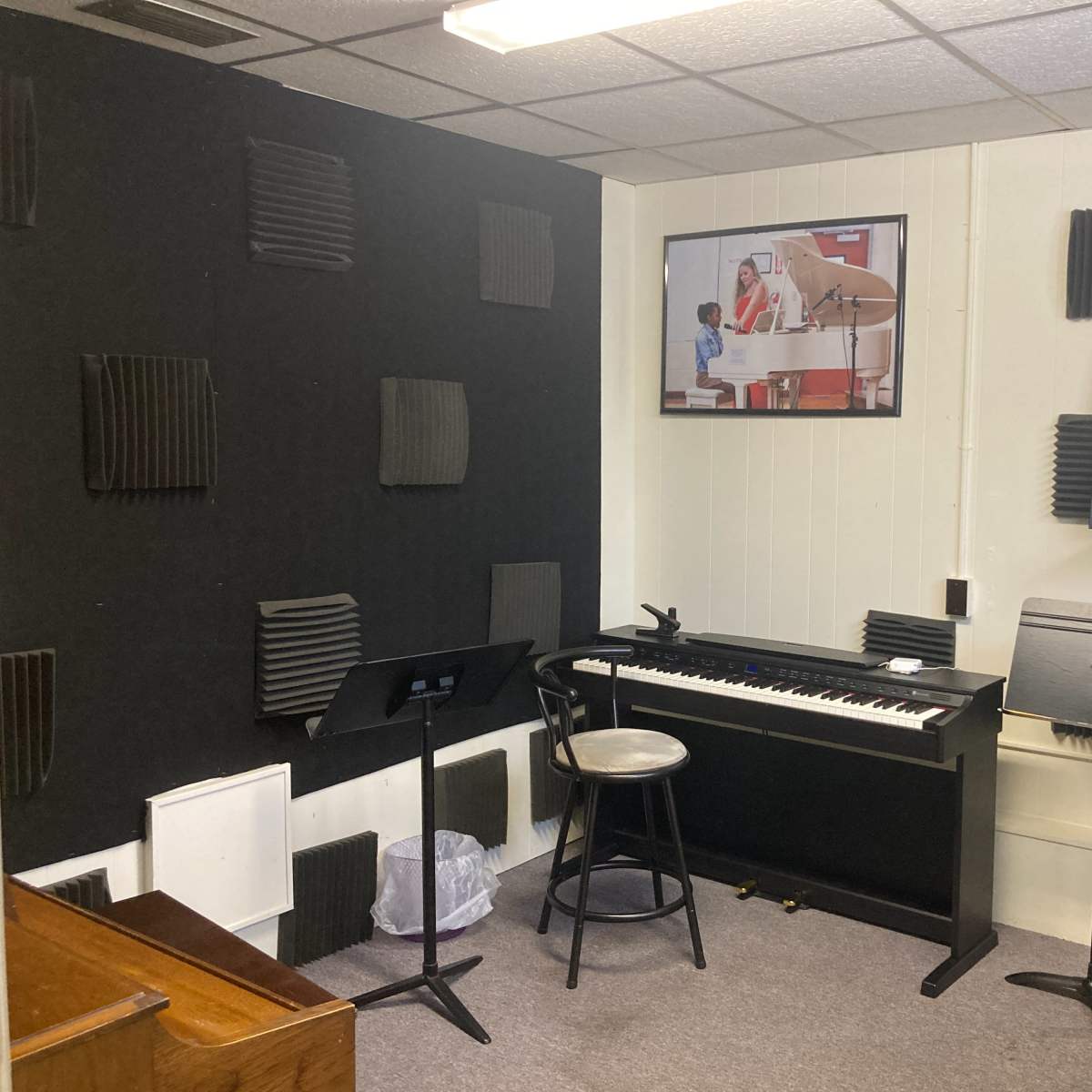 A music practice room equipped with a keyboard on a stand, a music stand, acoustic panels on the walls, and a fan in the corner.