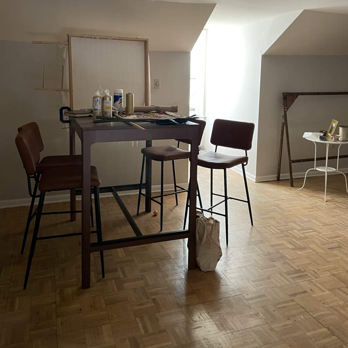 A spacious room with a high table and three chairs, illuminated by natural light from a window, along with an easel and a small table in the background.