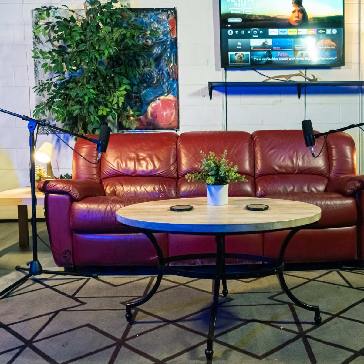 The scene features a red leather couch with two microphones on stands, a circular wooden coffee table with a small plant, and a television mounted on the wall above.