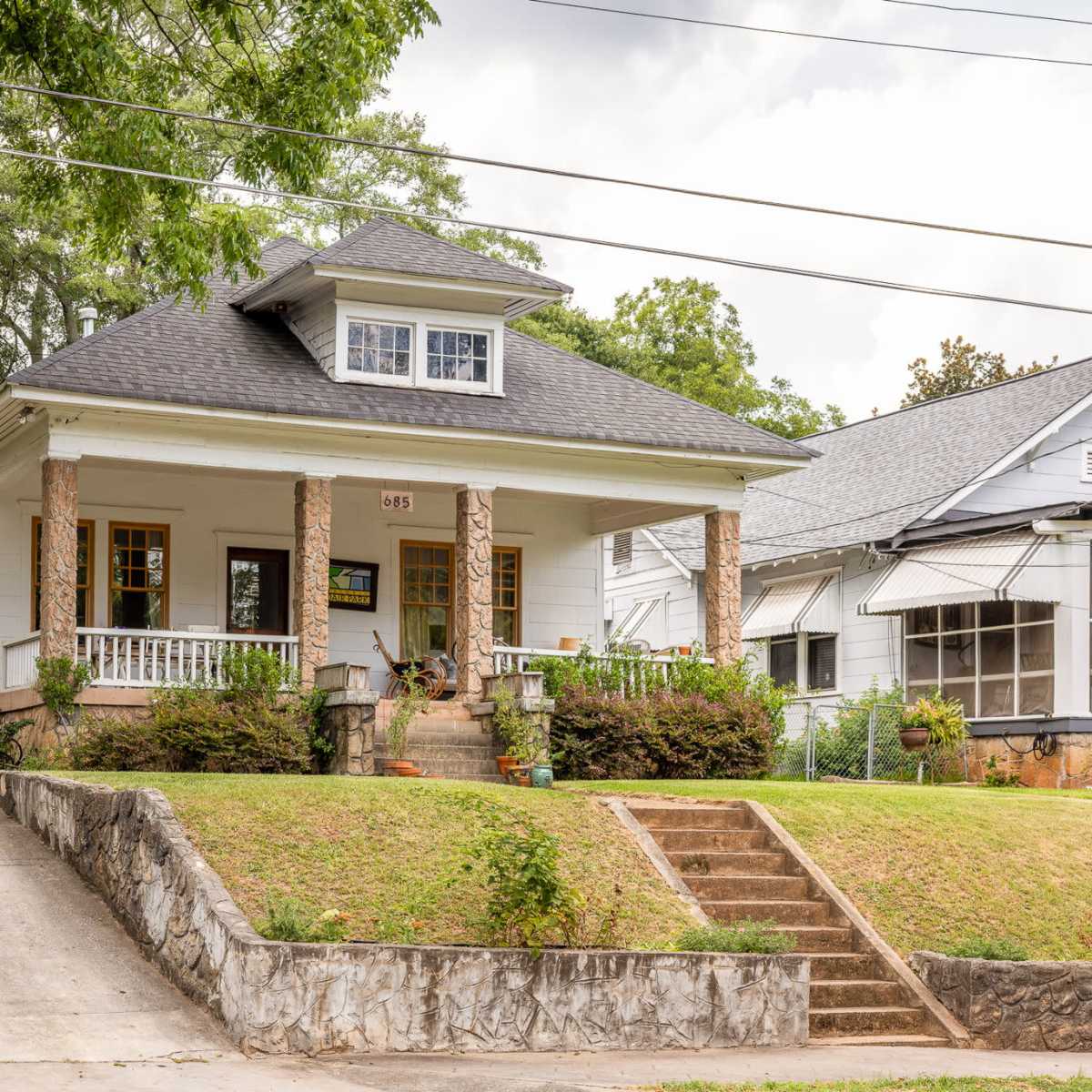 A single-story house with a gabled roof features a front porch supported by stone columns and is set on a grassy yard with a gravel driveway.