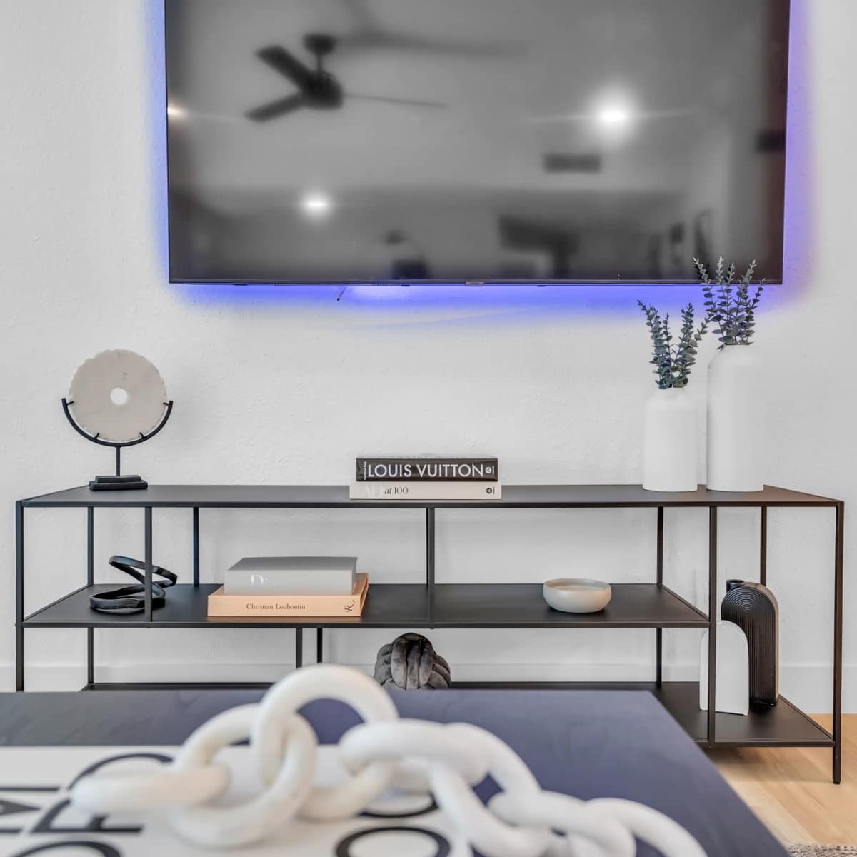 A modern living room features a sleek TV mounted on the wall above a black metal media console with decorative items and books.