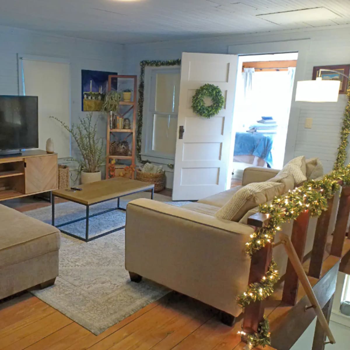 A living room with a television on one wall, a coffee table in the center, a couch, an ottoman, and a doorway leading to another room.