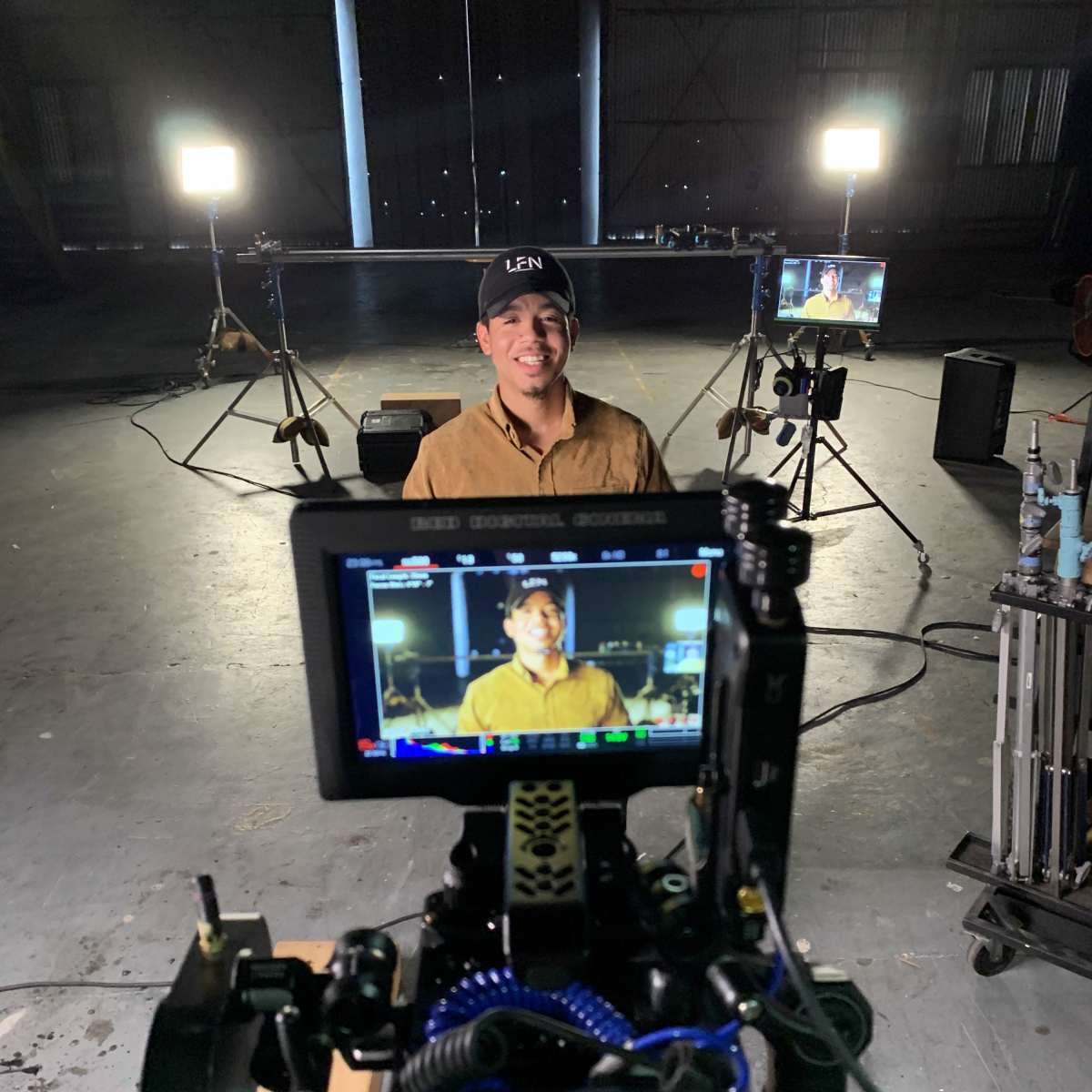 A man sits in front of a camera setup in a dimly lit studio, with lights positioned on either side and a monitor displaying his image.