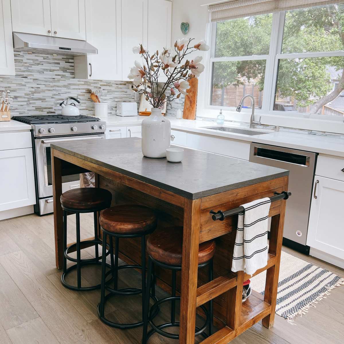 A modern kitchen features a wooden island with black stools, a decorative vase with branches, and white cabinetry.