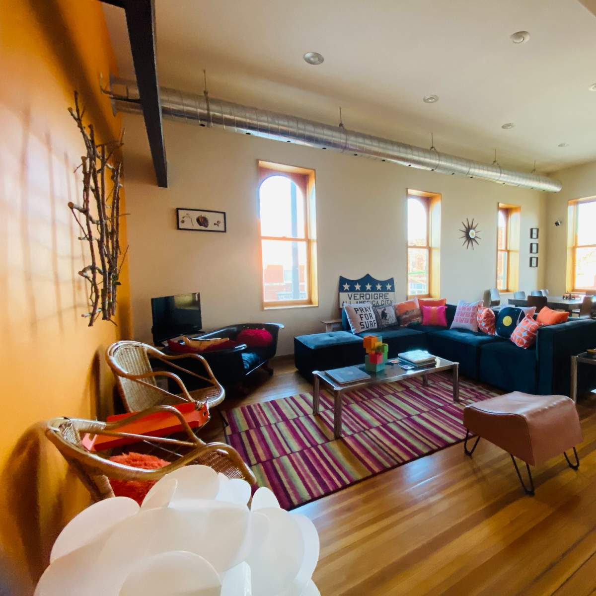A cozy living room features a large sectional sofa, colorful cushions, a coffee table, and large windows, with hardwood floors and an adjoining dining area.