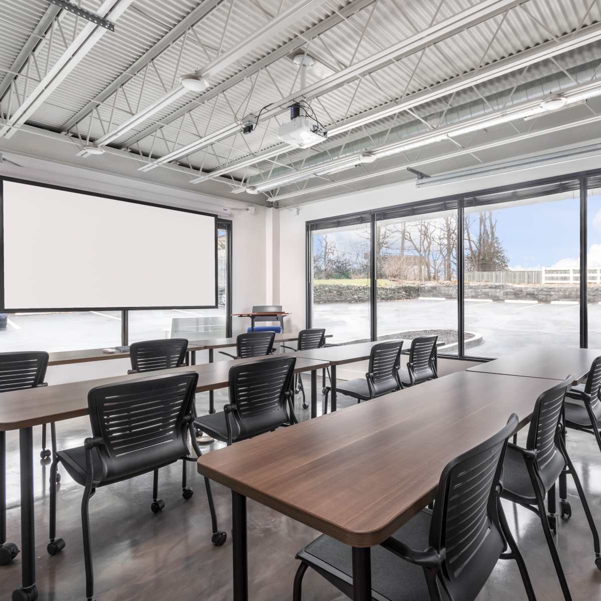 A modern classroom features multiple tables arranged in a U-shape, with black chairs, and large windows overlooking an outdoor area.