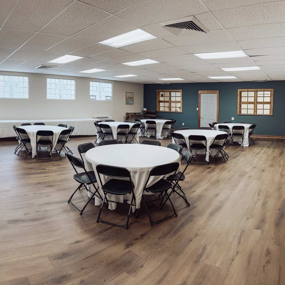 A spacious indoor venue features several round tables with white tablecloths and black chairs arranged on a polished wooden floor.