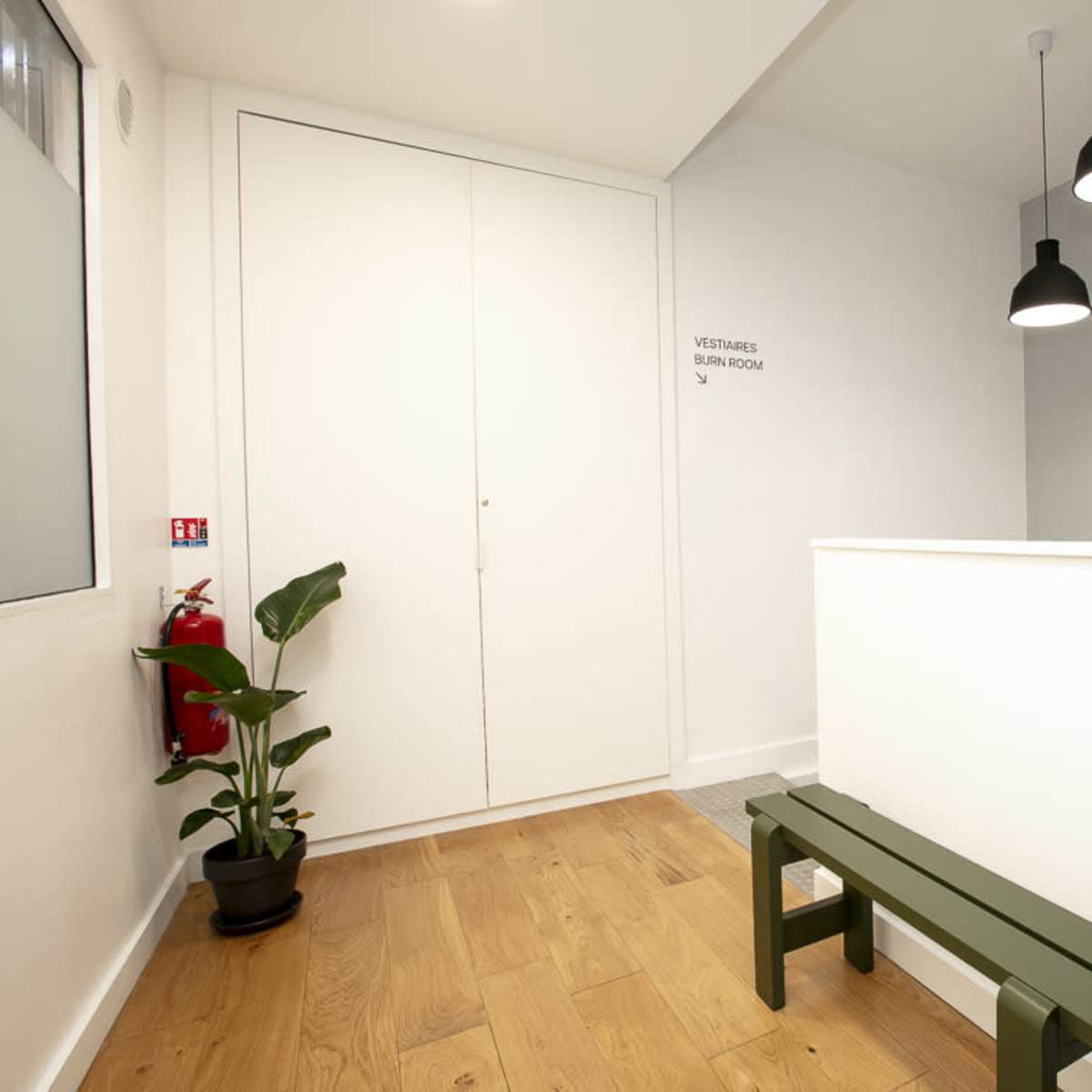 A bright waiting area with a bench, a water cooler, a fire extinguisher, and a potted plant against a light-colored wall.