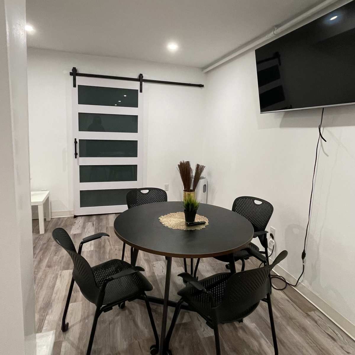 A modern room with a round black table and five black chairs, featuring a sliding glass door and a mounted television on the wall.