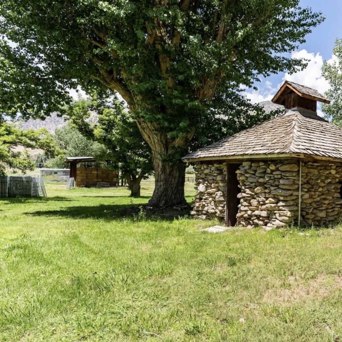 A stone building with a wooden roof stands in a grassy area surrounded by trees.