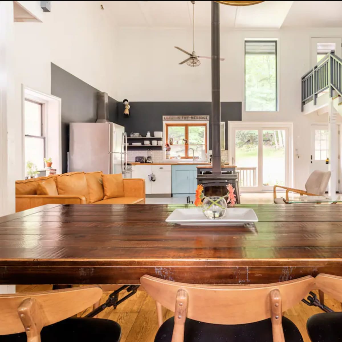 A wooden dining table with several chairs is foregrounded in a spacious kitchen and living area featuring a staircase and large windows.