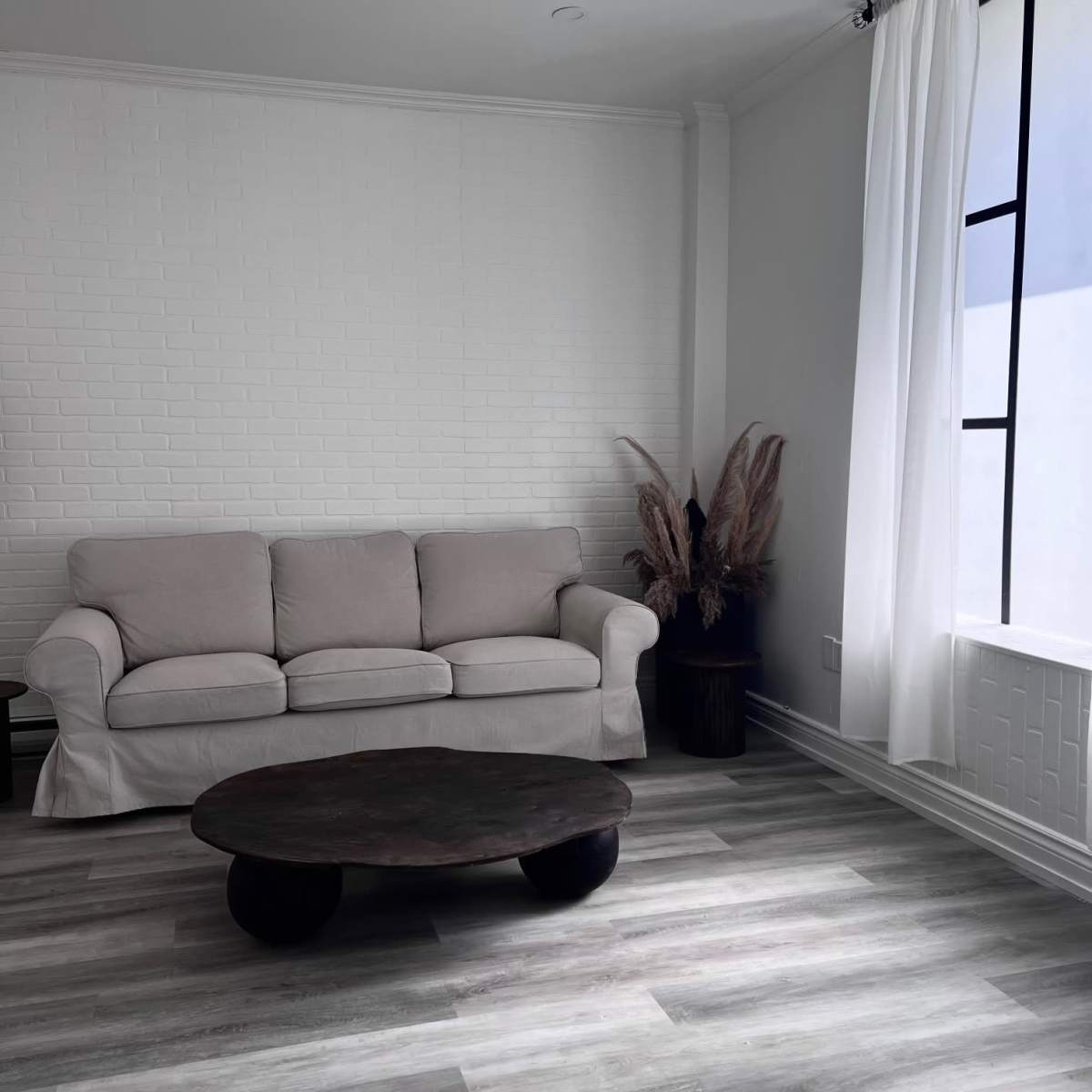A simple living room with a light-colored sofa, a round wooden coffee table, and a window adorned with sheer curtains.