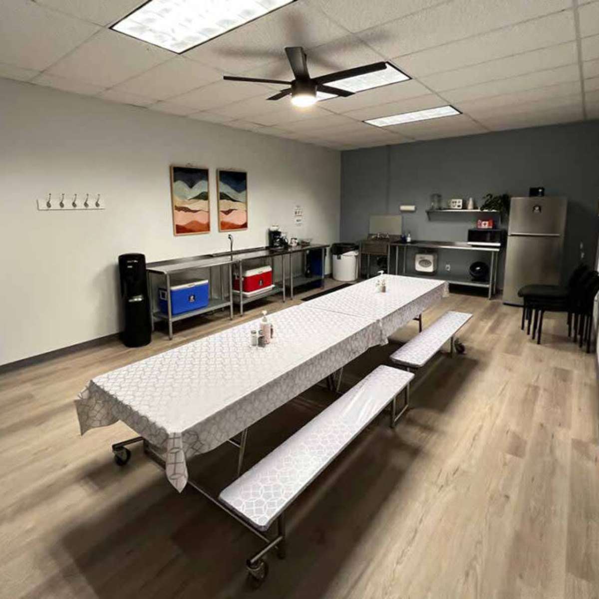 A clean, minimally furnished break room features a long table with benches, a kitchen area, and several artworks on the walls.