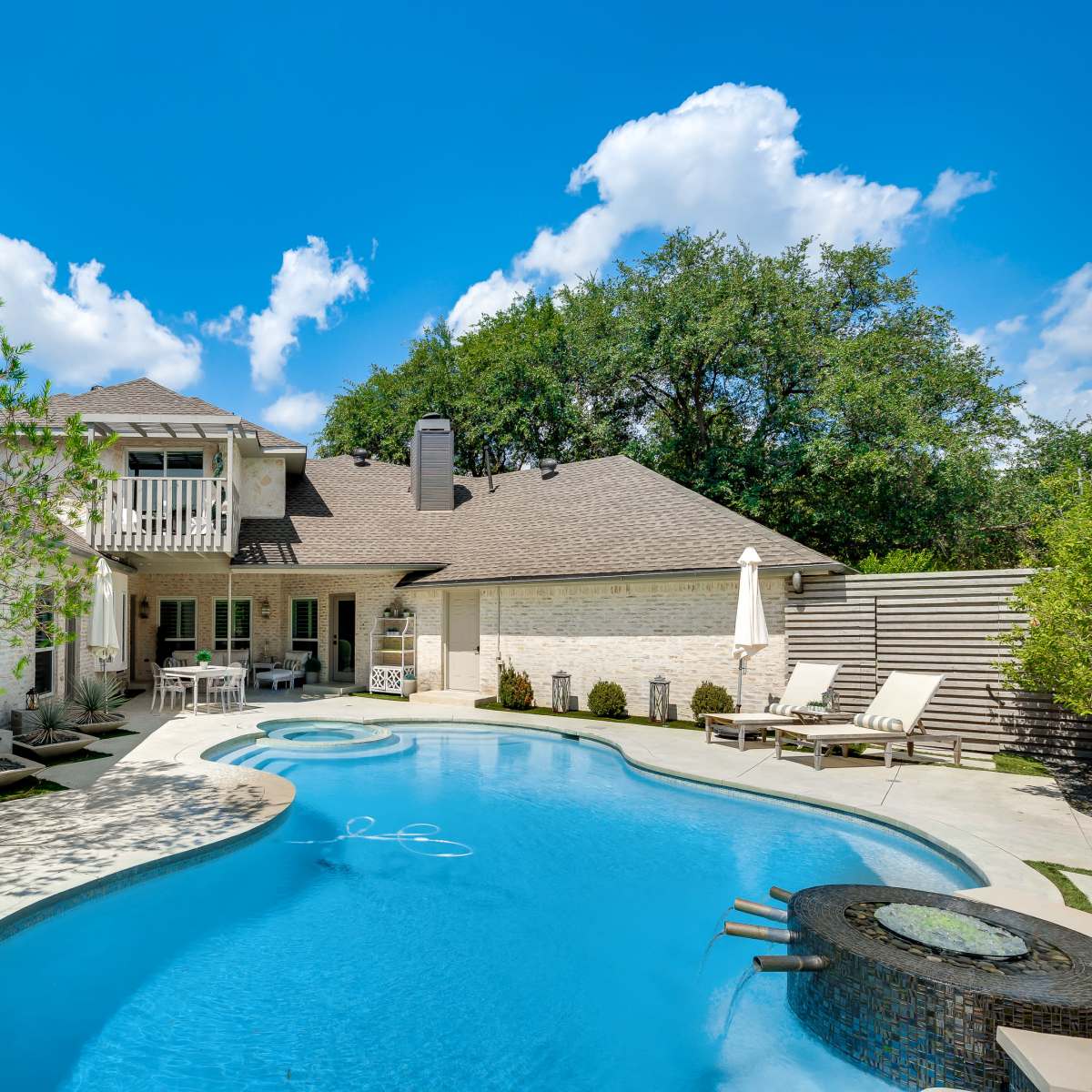 A backyard with a swimming pool, a patio area with seating, and a two-story house surrounded by greenery.