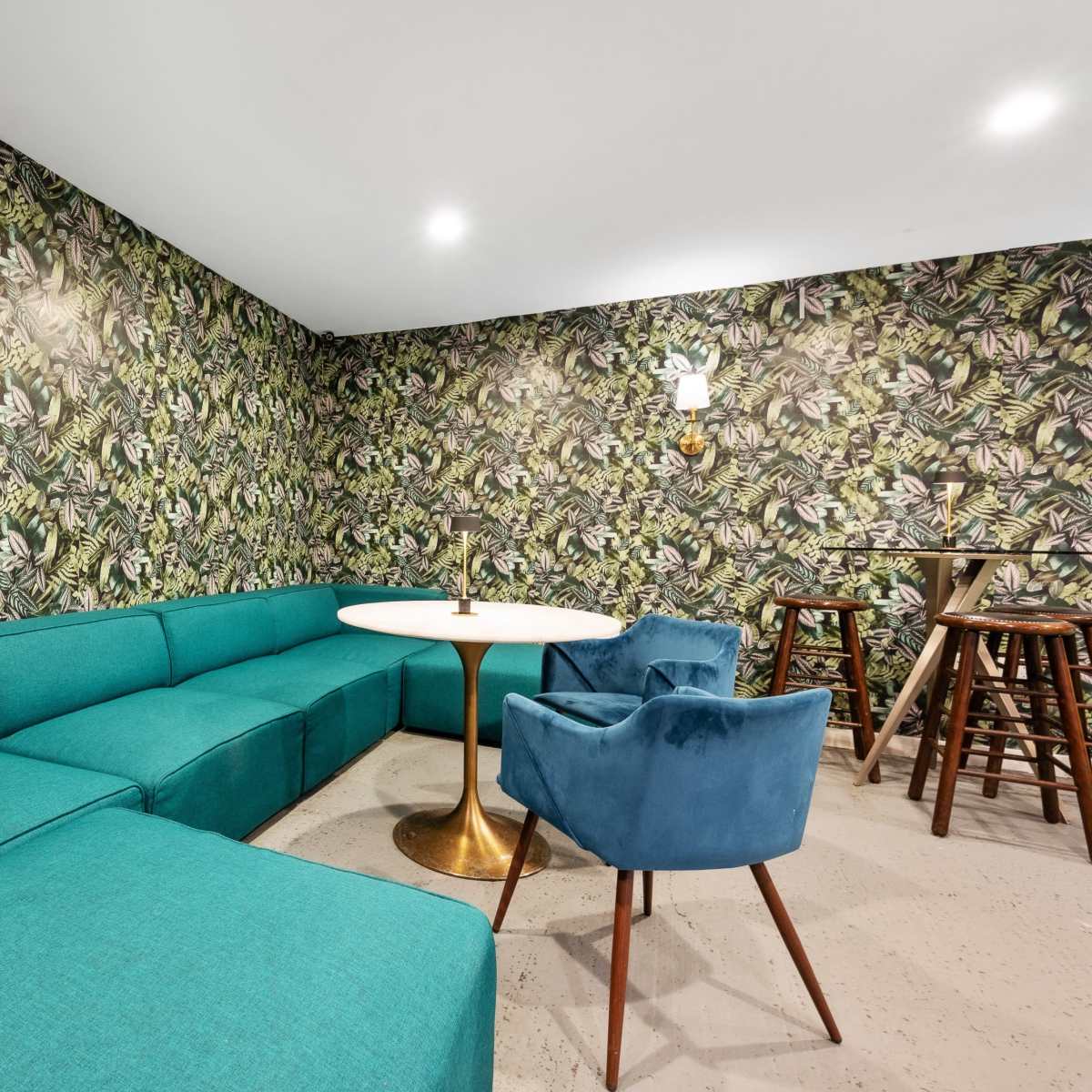 A seating area with a green sectional couch, a round table, and blue chairs, all set against a patterned wallpaper featuring tropical foliage.