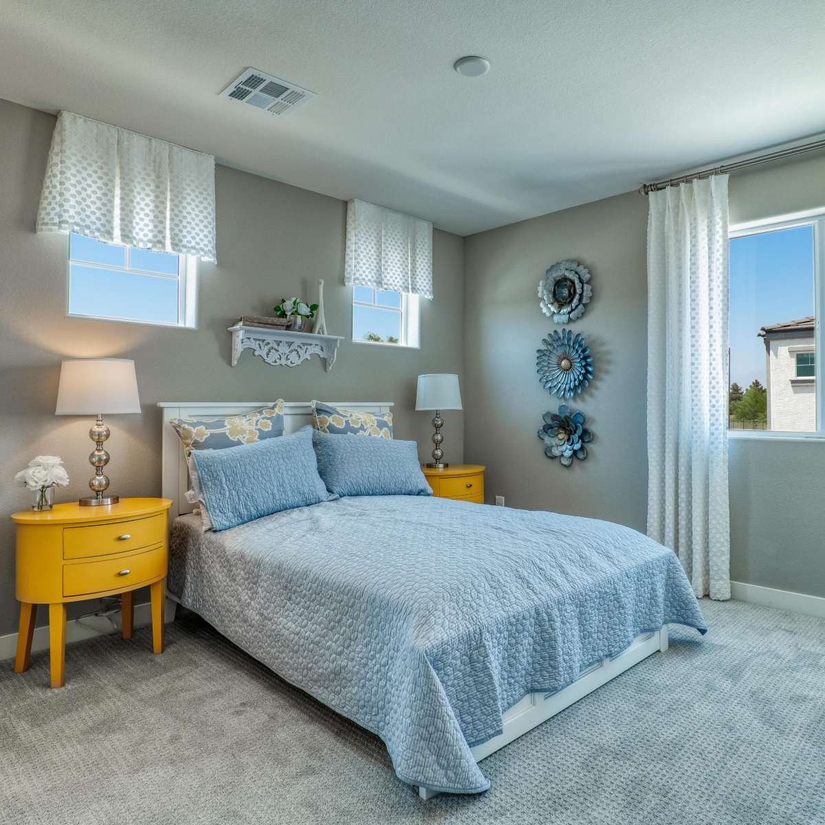 A neatly arranged bedroom features a bed with a light blue coverlet, two bedside tables, and wall decor including circular mirrors and window treatments.