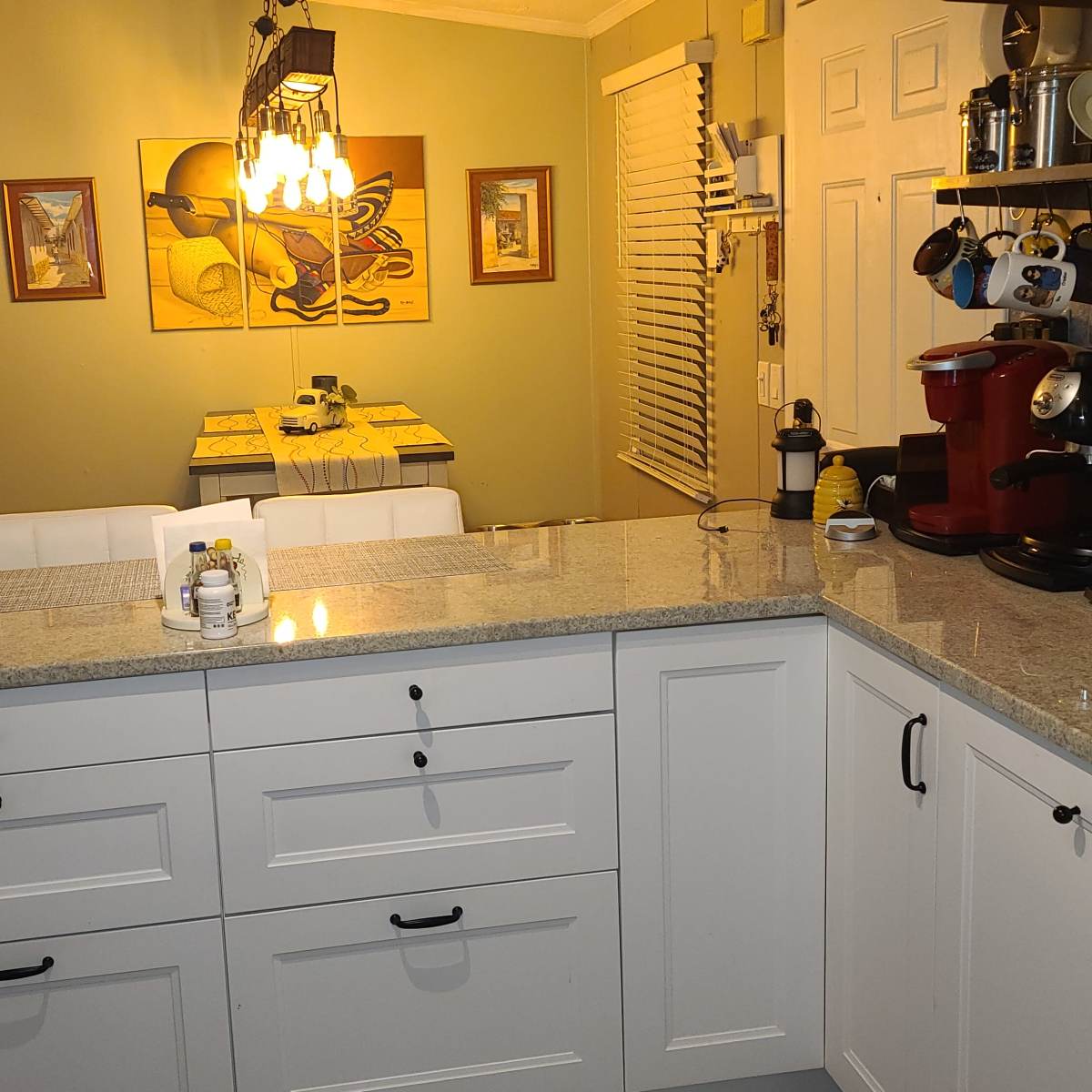 A spacious kitchen features a granite countertop with white cabinetry, a coffee maker, and a dining area visible in the background.