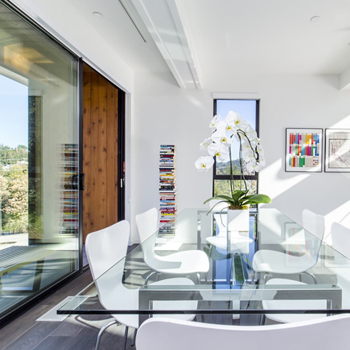 A modern dining area features a glass table surrounded by white chairs, with a large orchid centerpiece and a wall-mounted television.