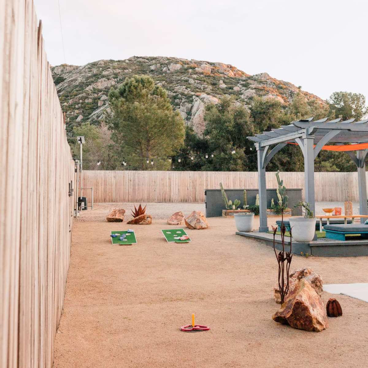 A landscaped outdoor area features a mini-golf setup with green mats and rocks, framed by a wooden fence and backed by a rocky hillside.