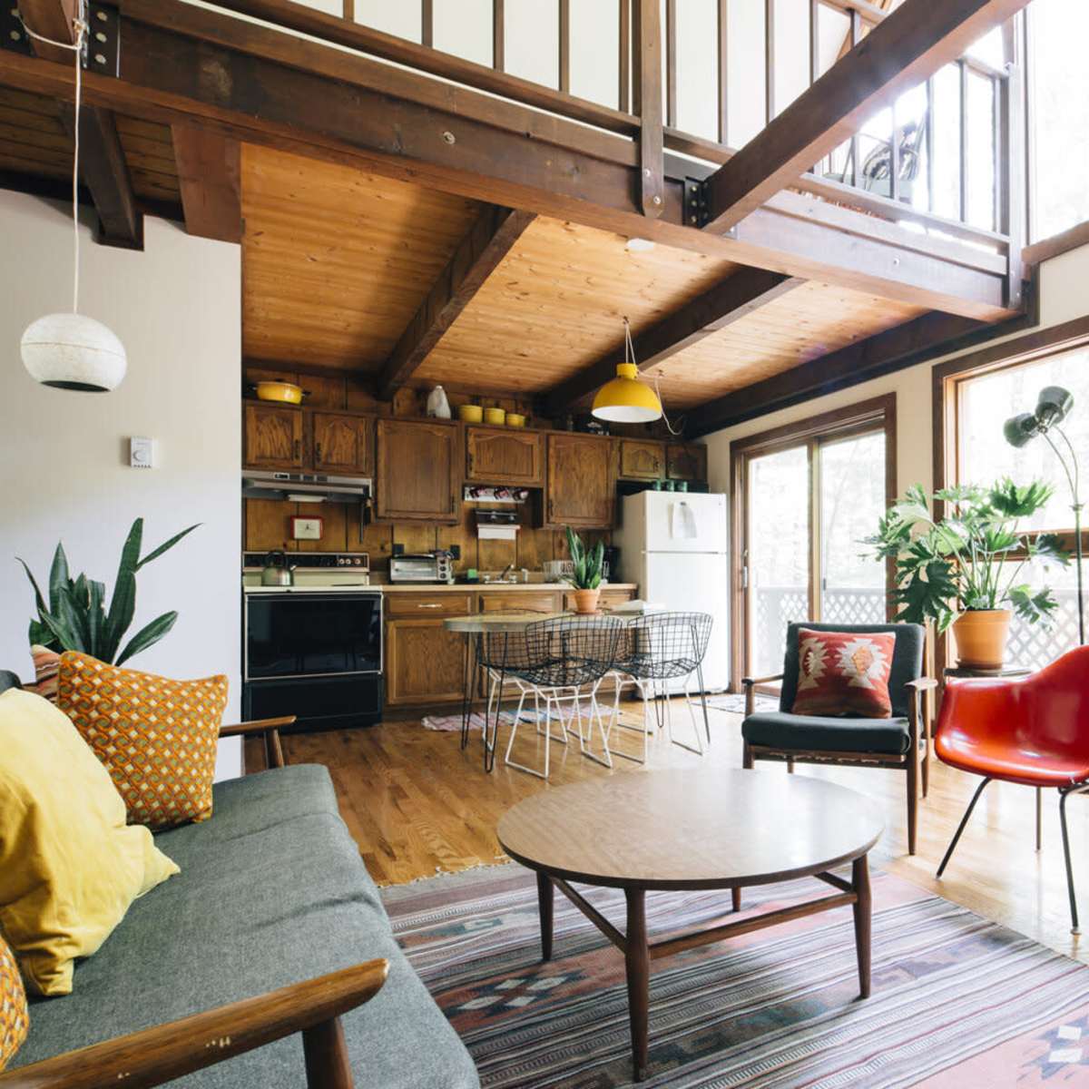 A spacious living area with wooden beams, a modern kitchen in the background, and large windows allowing natural light to enter.