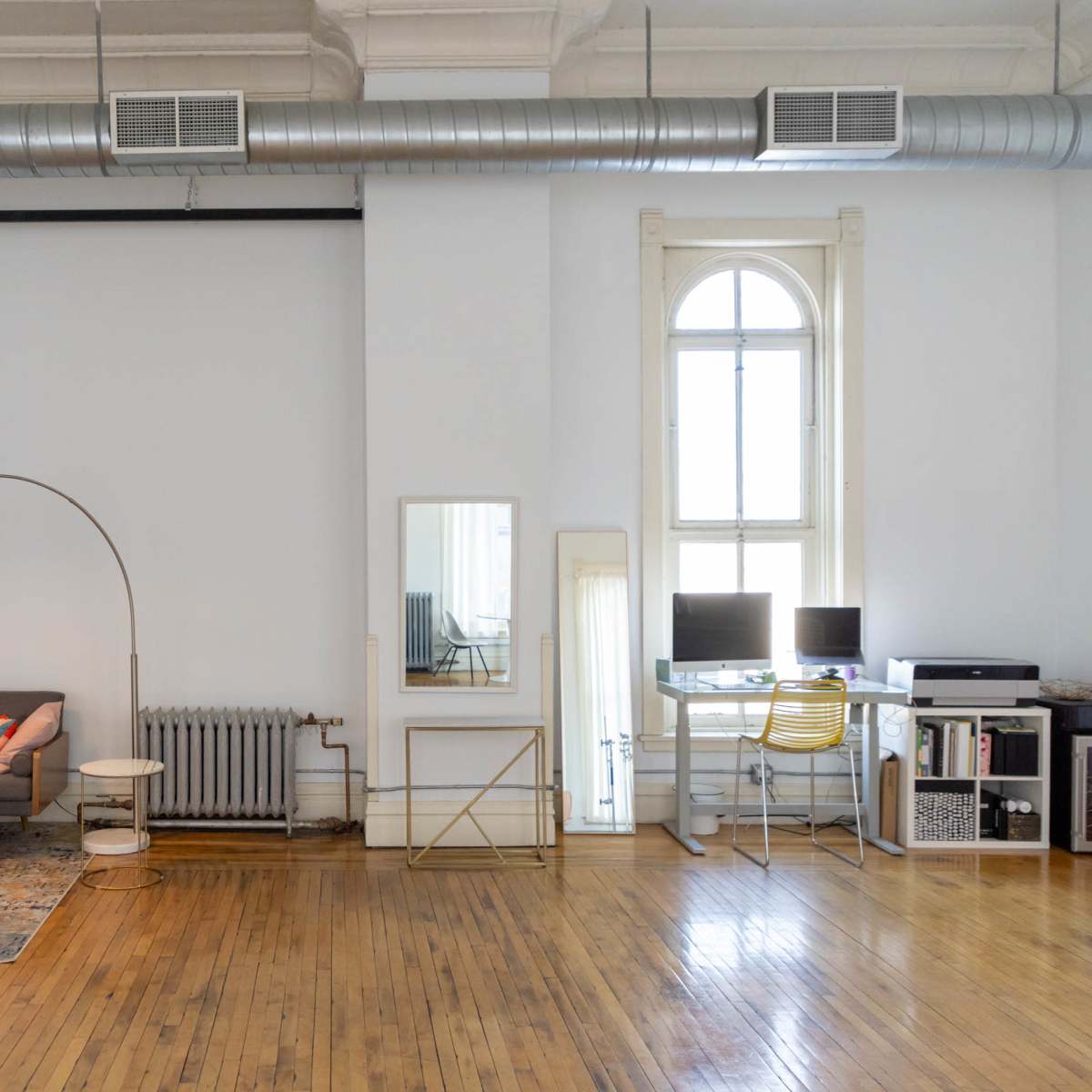 A spacious living area with a gray sofa, wooden coffee table, and desk setup, featuring large windows and wooden flooring.