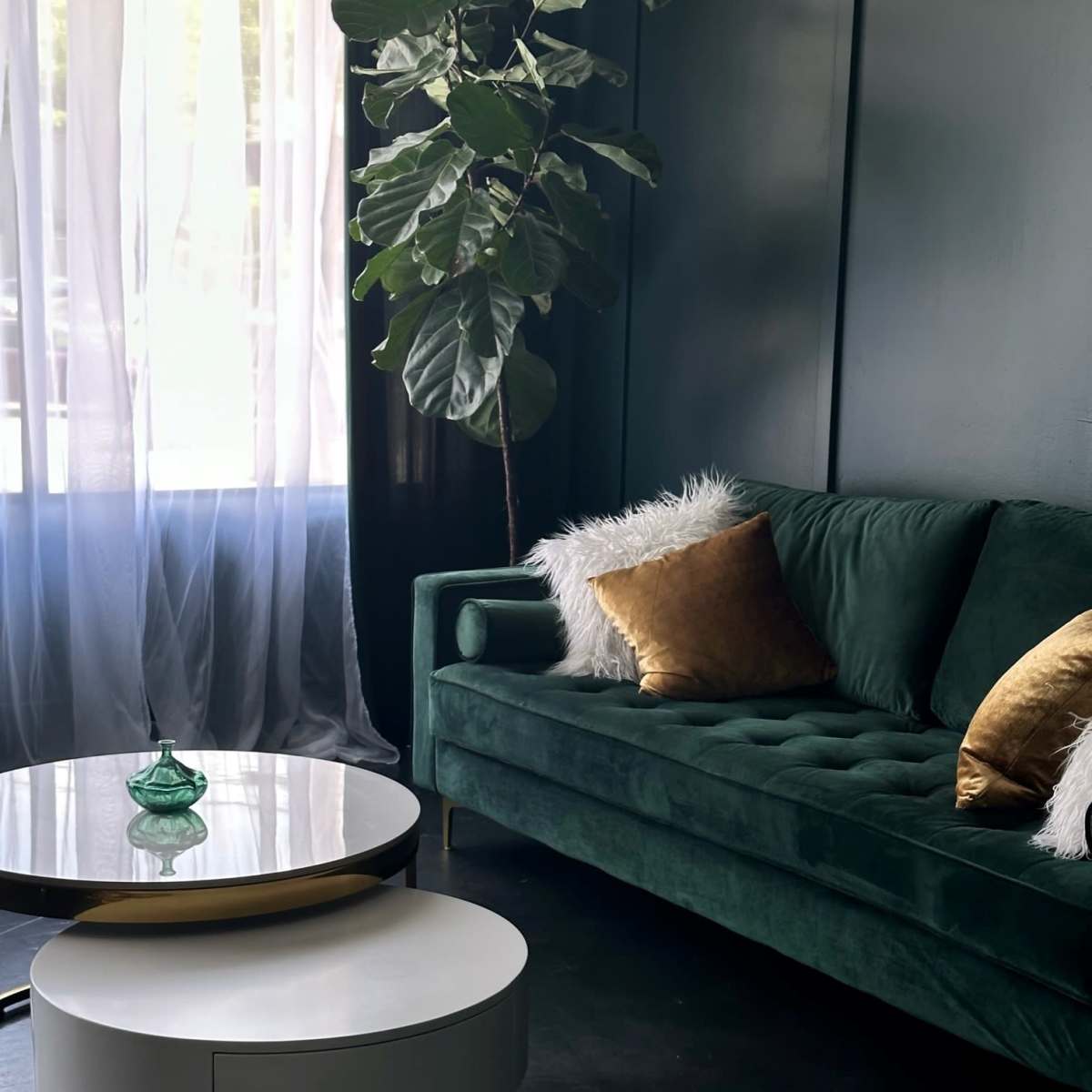 The scene features a green velvet sofa with gold and white decorative pillows, a circular coffee table in white and gold, and a potted plant beside a window with sheer curtains.