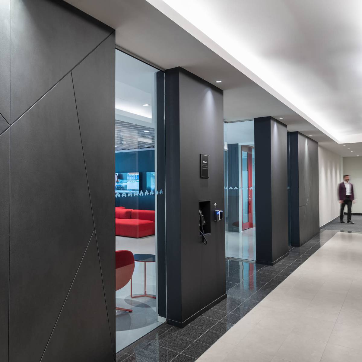 A modern office hallway features a textured black wall leading to a glass-fronted meeting room with red seating.