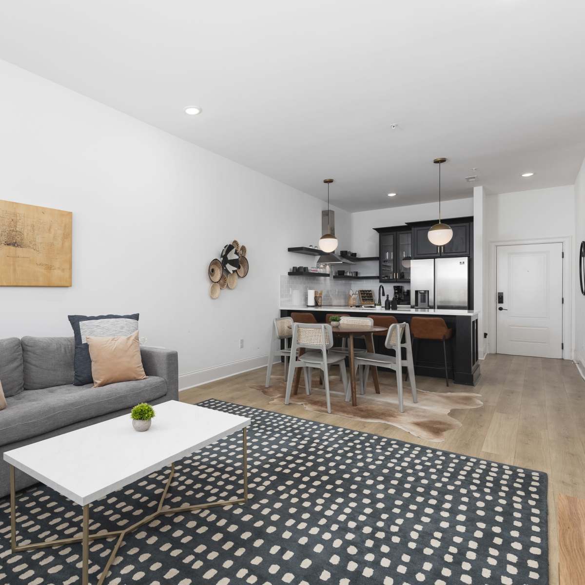 A modern living room features a gray sofa with decorative pillows, a white coffee table, and a kitchen area in the background with dark cabinetry and bar seating.