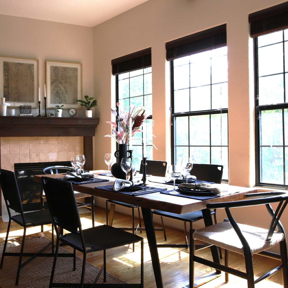 A dining room features a wooden table set for a meal, surrounded by black chairs, with large windows allowing natural light and a fireplace along one wall.