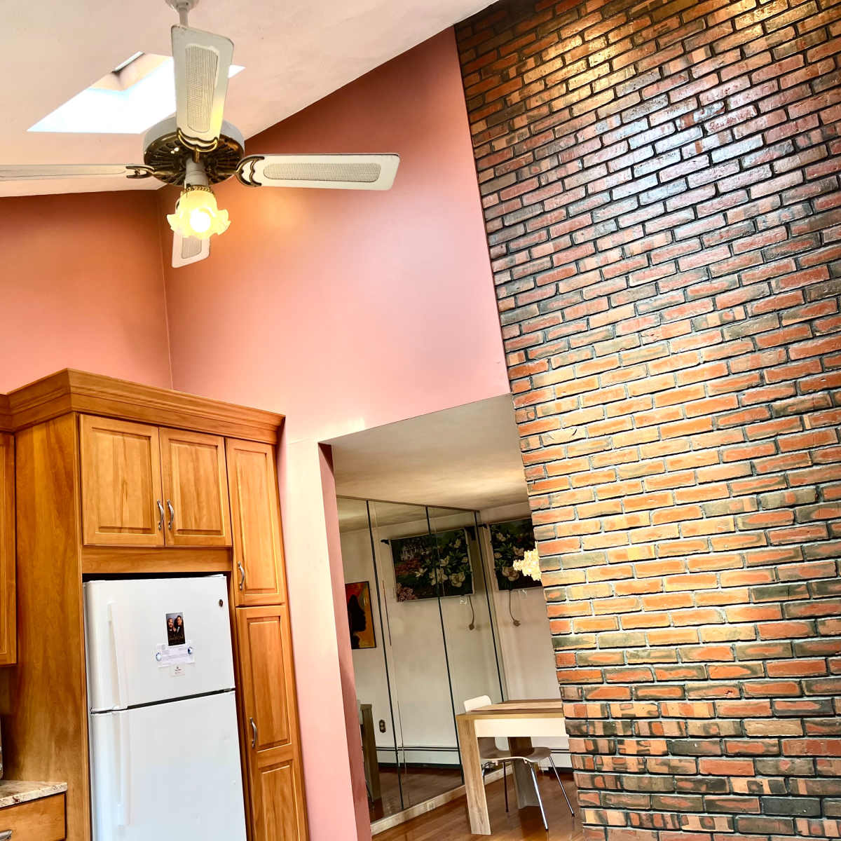 A kitchen with wooden cabinets, white appliances, a ceiling fan, and a brick wall adjacent to a dining area with a table.