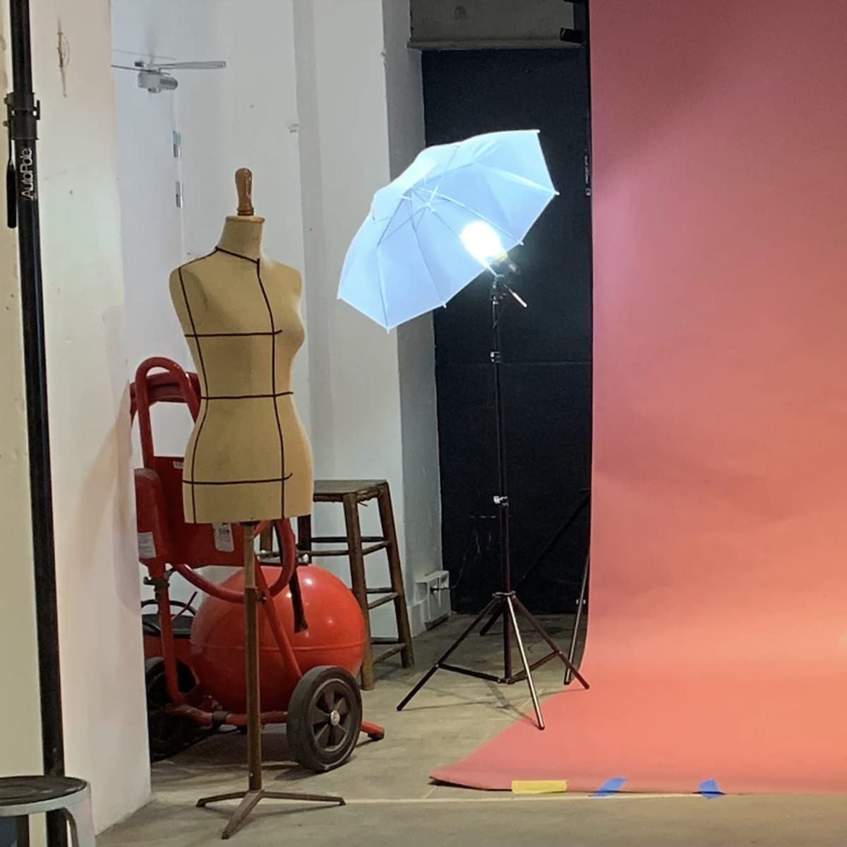 A photography studio setup with a pink backdrop, two softbox lights, and various equipment including a dress form and a chair.