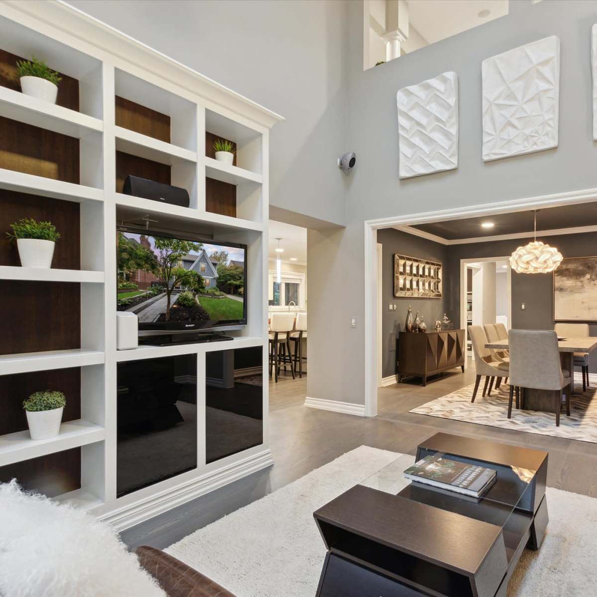 A modern living room features a built-in shelving unit with a TV, a sectional sofa, and a dining area visible in the background.